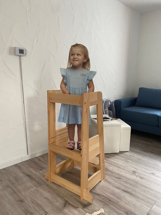 A smiling child in a blue dress standing on a wooden step stool in the living room.