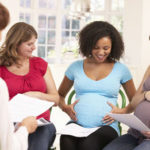 Four women are seated, three of whom are visibly pregnant, holding documents, in a bright room.