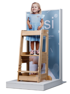 Child model standing on wooden step stool in front of promotional backdrop.