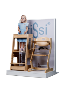 Child in blue dress standing on wooden step stool beside adjustable high chair.