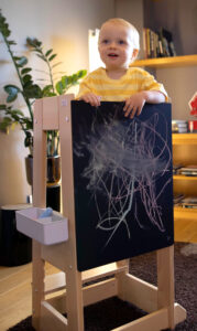 Toddler in yellow shirt standing at chalkboard with colorful scribbles, indoor setting.