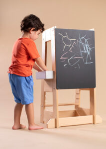 Child in orange shirt drawing with chalk on a blackboard easel indoors.